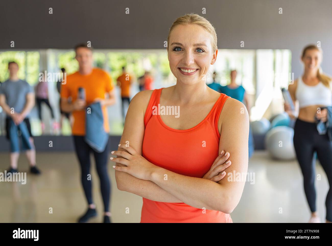 Donna sicura di sé con canotta arancione in piedi con le braccia incrociate in palestra. Immagine del concetto di lavoro di squadra Foto Stock