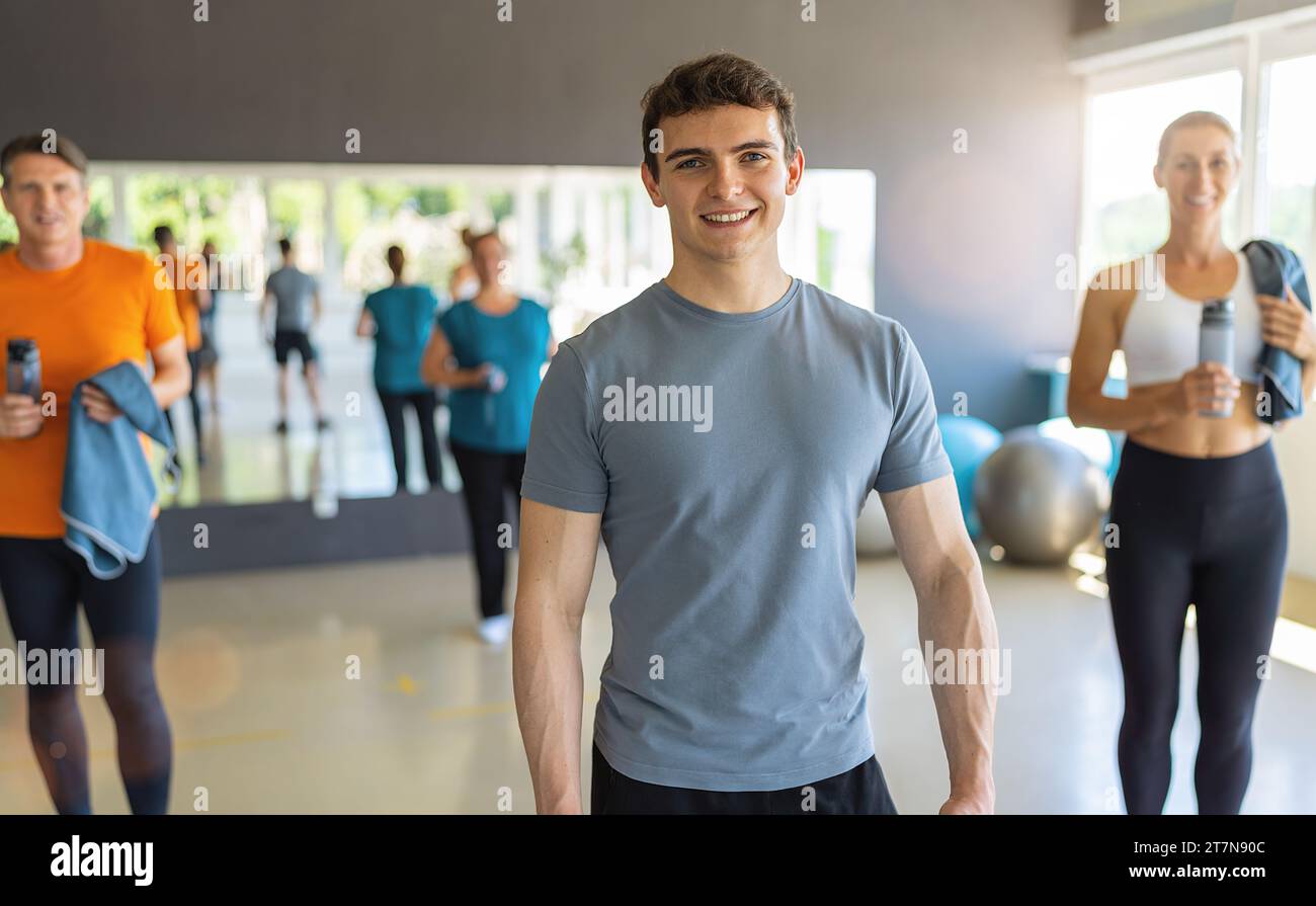 Giovane uomo con t-shirt grigia sorridente in palestra con altre persone sullo sfondo. Immagine del concetto di lavoro di squadra Foto Stock
