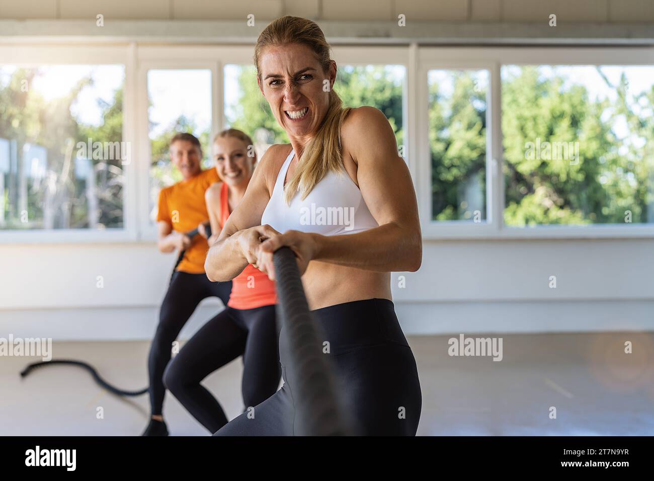 la donna tira su una corda con espressioni facciali espressive come una gara di tiro di guerra in un gruppo come esercizio di teambuilding. Palestra per esercizi di fitness conc Foto Stock