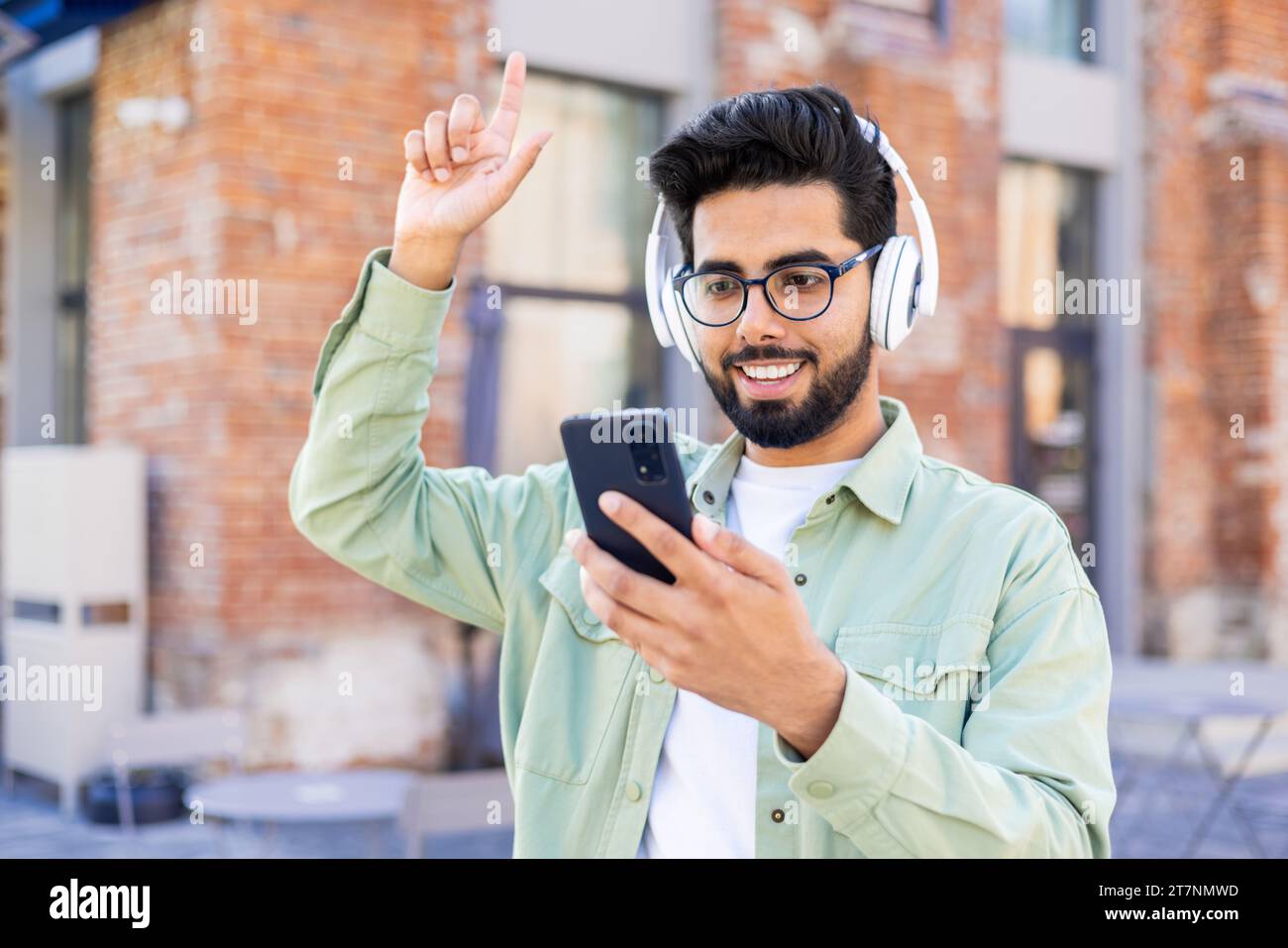 Un giovane con le cuffie cammina per la città, gioioso, sorridente, ascolta musica online, utilizzando un'applicazione al telefono. Foto Stock