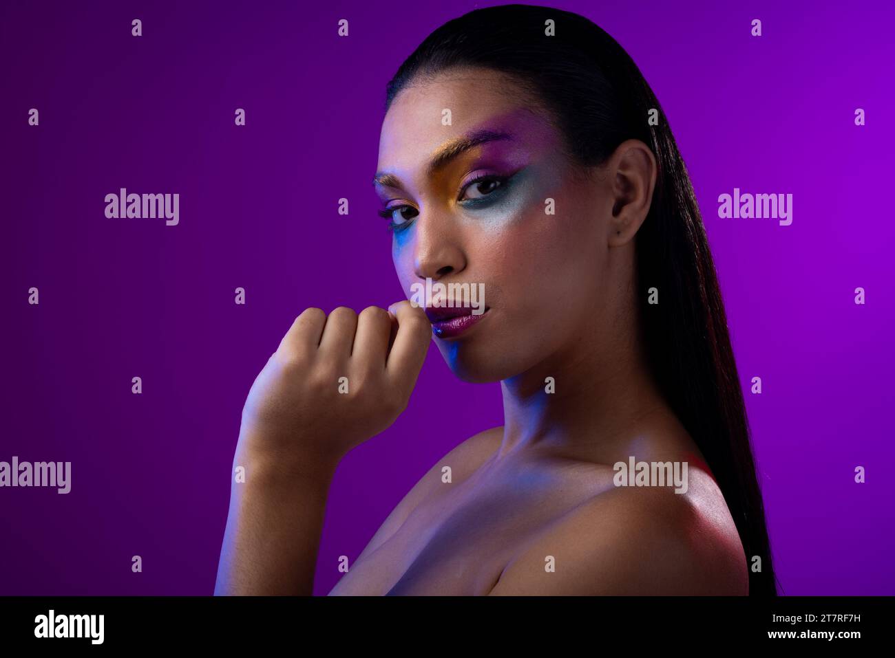 Donna birazziale con capelli scuri, mano a bocca, labbra rosa, trucco con ombre oculari su sfondo viola Foto Stock