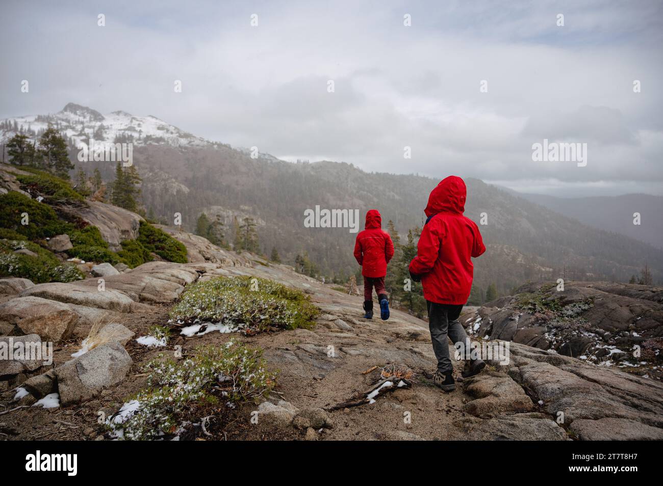 Due adolescenti camminano in rosso lungo un masso di granito con montagne innevate Foto Stock