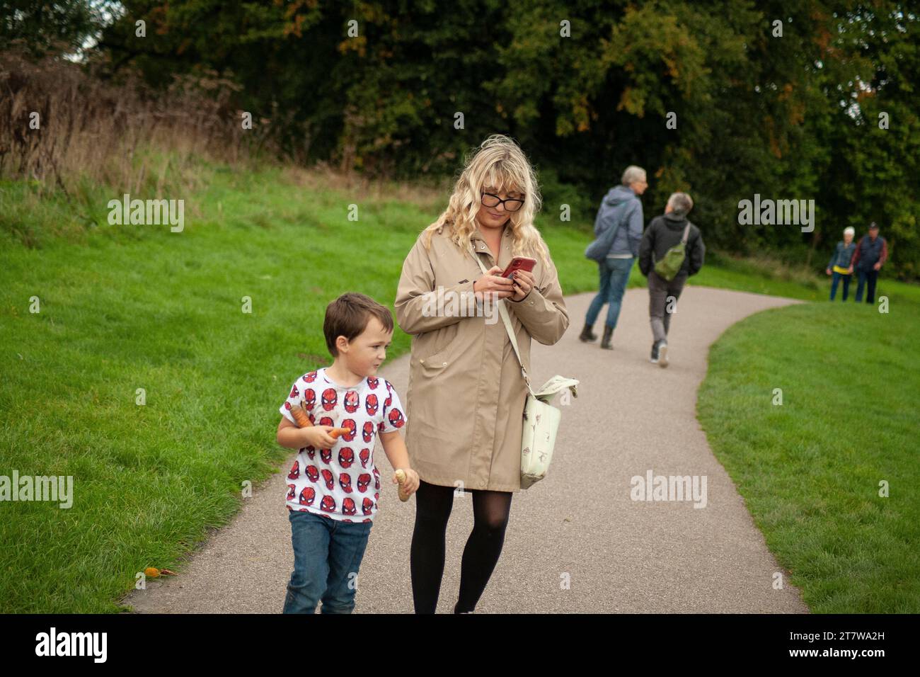 Una giovane madre passeggia in un parco con il suo piccolo, navigando nella bellezza del parco mentre tenta di usare il suo cellulare. genitori contemporanei Foto Stock