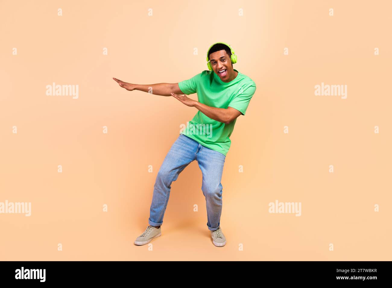 Foto a lunghezza intera di un uomo positivo estatico indossa eleganti jeans t-shirt Ascolta musin in cuffie che ballano isolate su uno sfondo di colore beige Foto Stock