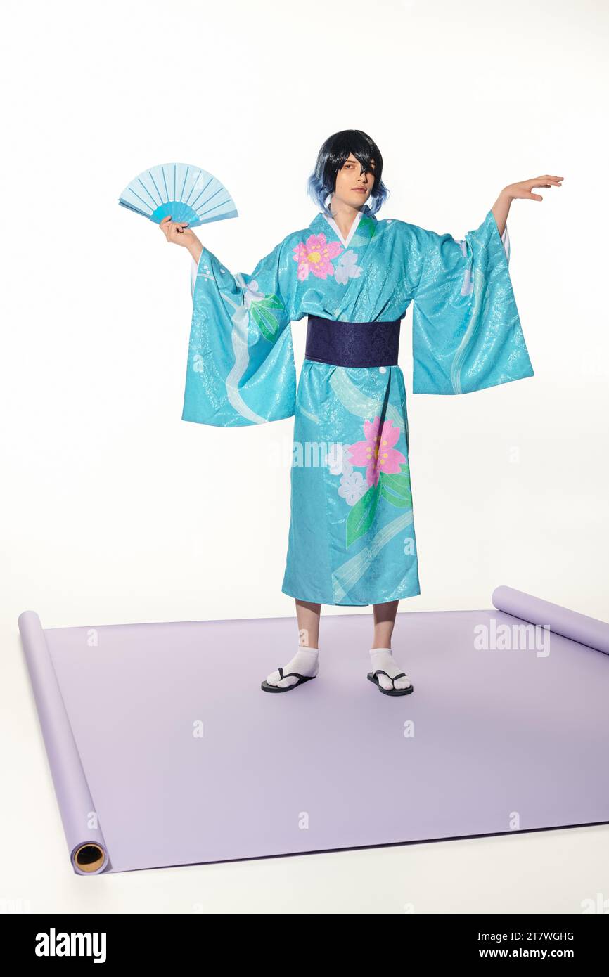 uomo espressivo in kimono blu e parrucca in posa con ventola a mano su tappeto viola e sfondo bianco Foto Stock