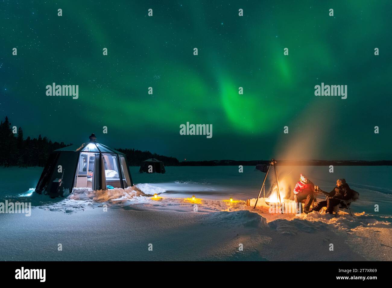 Vista notturna di due persone e cena all'aperto intorno a un falò vicino all'igloo di vetro illuminato con l'aurora boreale Foto Stock