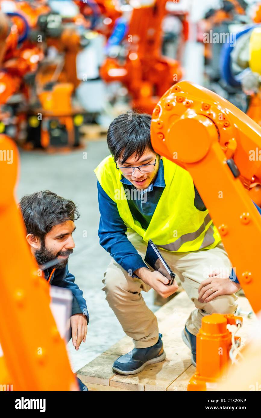 Foto verticale di due giovani ingegneri multietnici che esaminano il funzionamento dei bracci robotici nella fabbrica Foto Stock