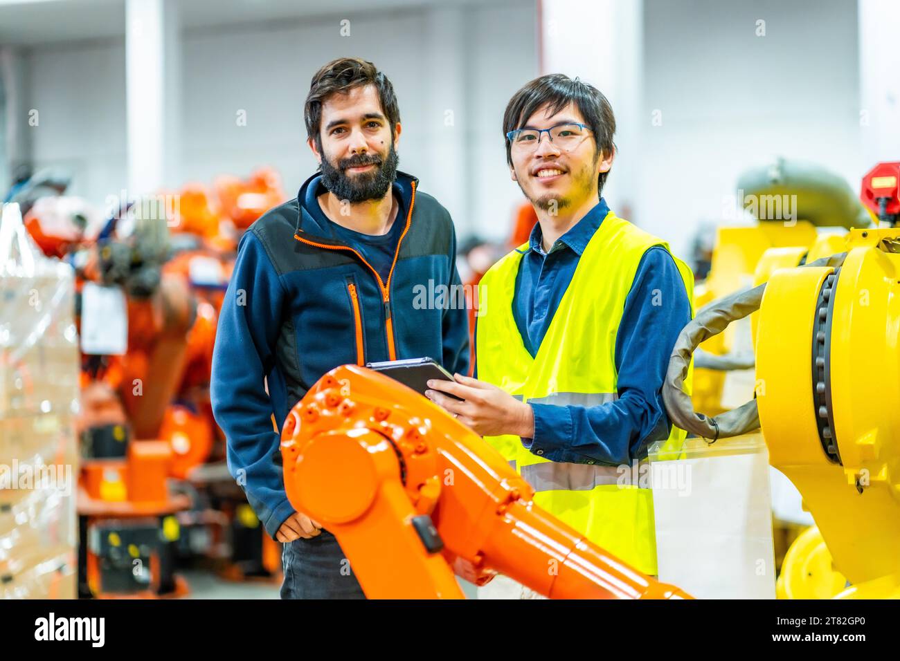 Ritratto di due giovani ingegneri multietnici in un'industria robotica industriale che guardano la macchina fotografica e sorridono Foto Stock