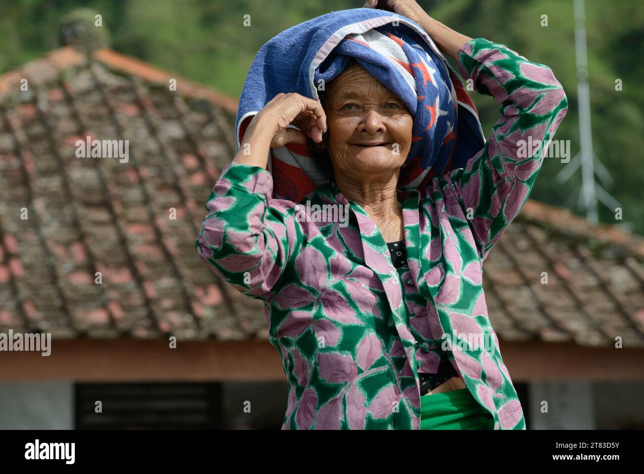 Una donna anziana balinese, il suo volto impreziosito dalla saggezza degli anni, si erge orgogliosamente in abiti tradizionali sullo sfondo della sua casa di villaggio Foto Stock