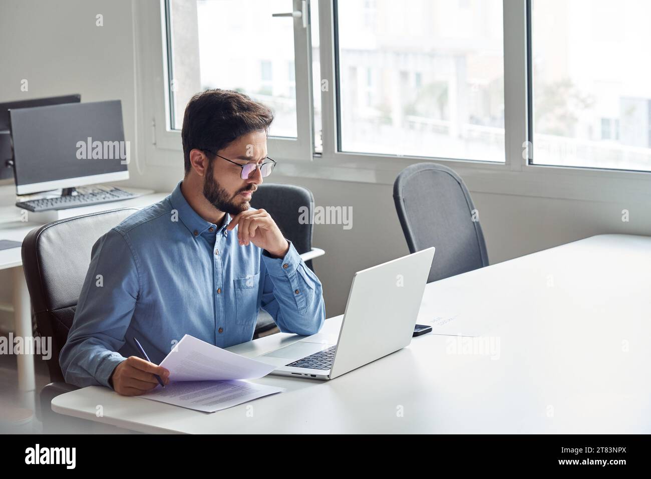 Giovane uomo d'affari impegnato che lavora su un computer portatile con documenti in mano. Foto Stock