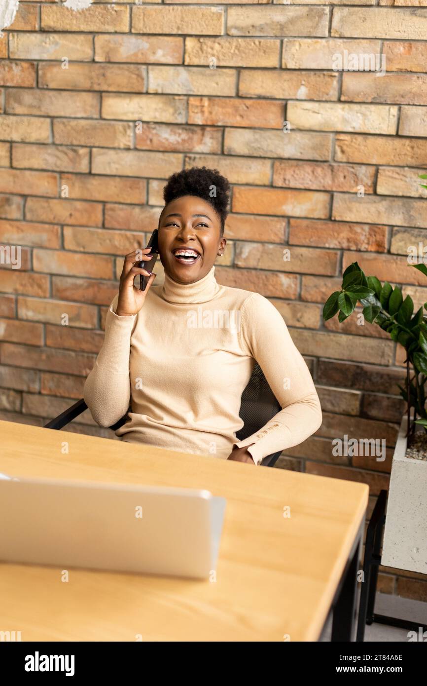 Una bella giovane donna d'affari afroamericana che usa il telefono cellulare seduto accanto al muro in mattoni nell'ufficio in stile industriale Foto Stock