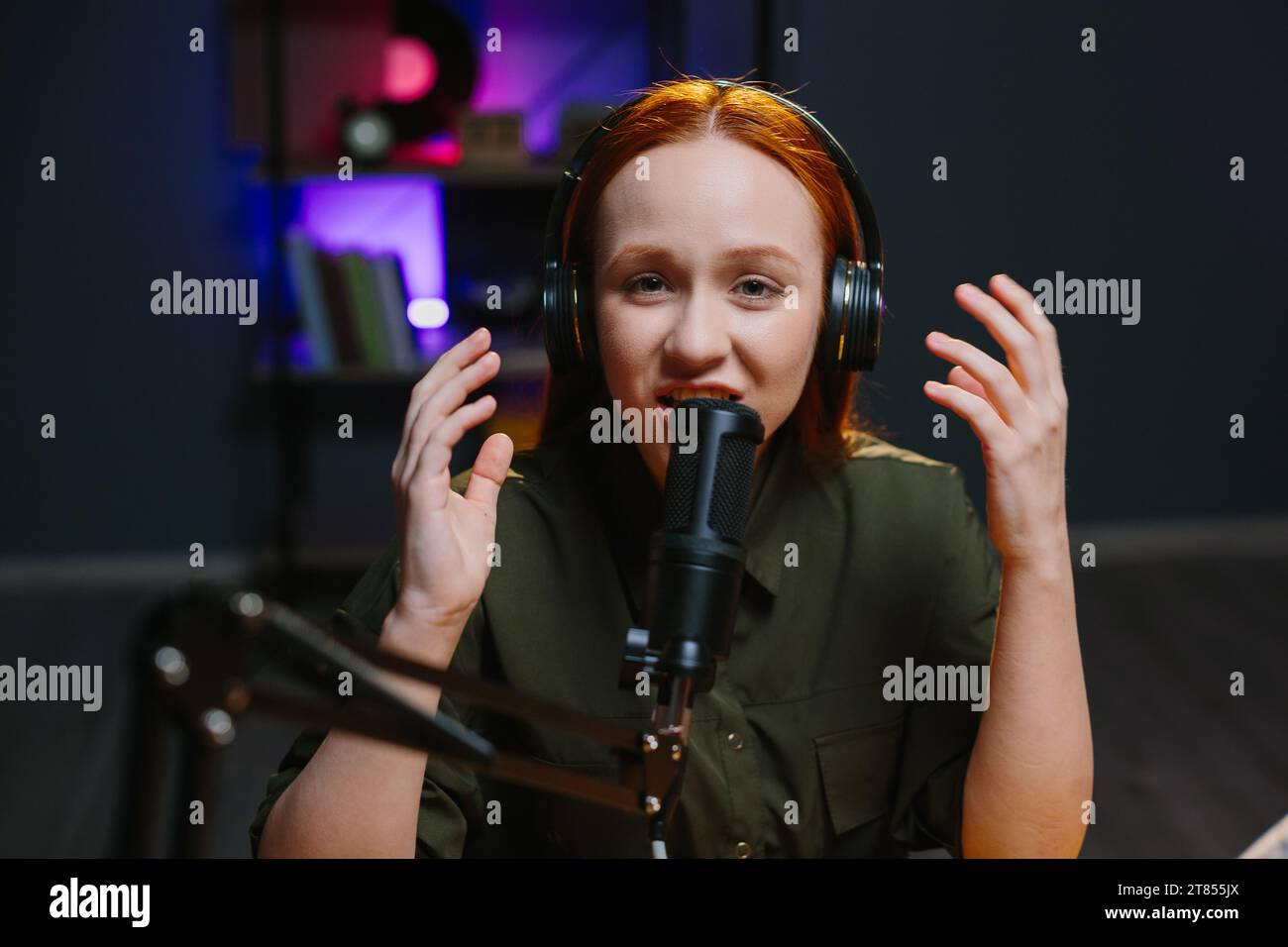 Una ragazza con le cuffie parla in modo espressivo nel microfono mentre registra un nuovo episodio del blog. Foto Stock