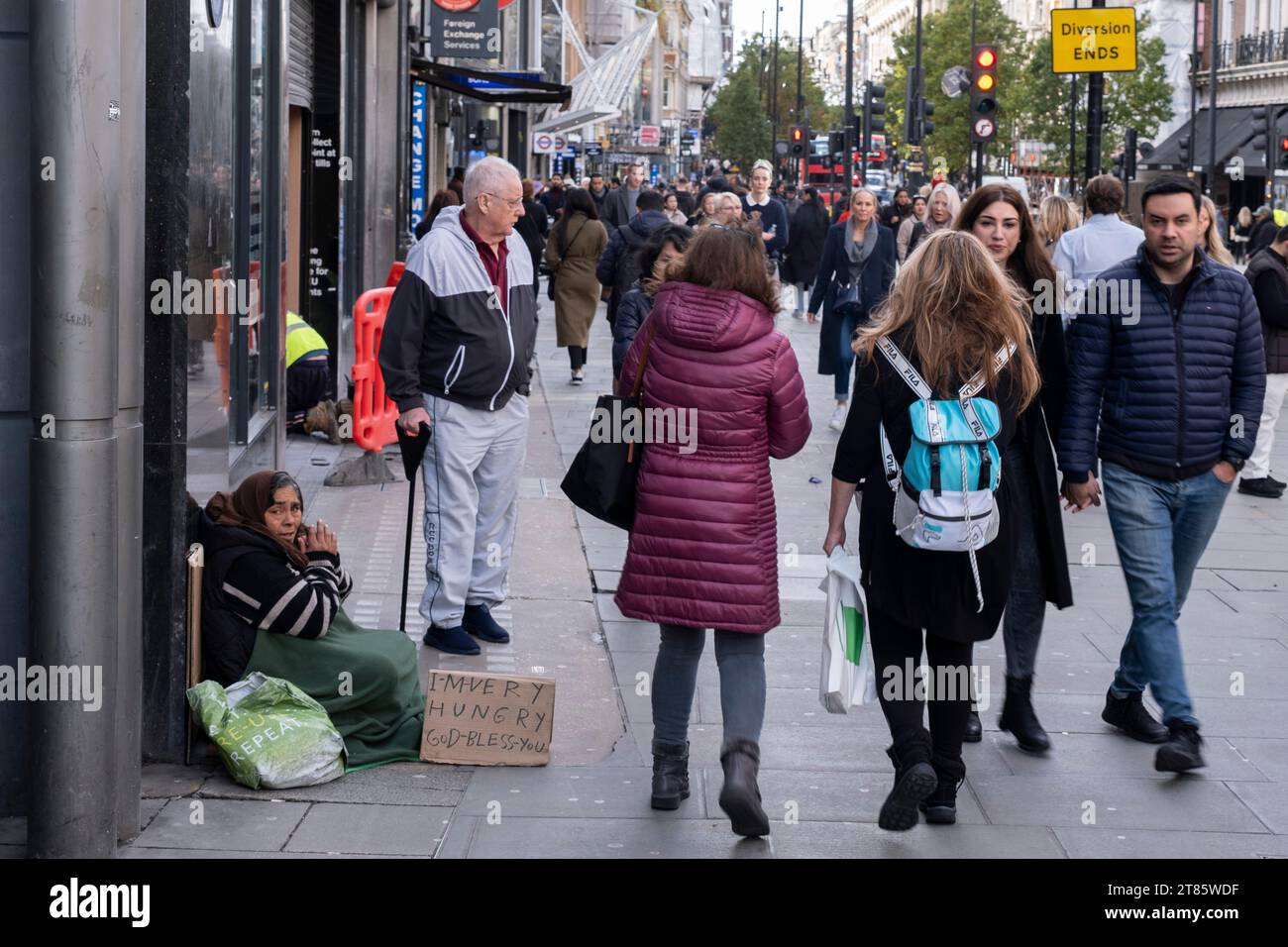 Mentre gli acquirenti e i visitatori passano da Oxford Street, una donna siede sul marciapiede a chiedere soldi il 13 novembre 2023 a Londra, Regno Unito. La scena illustra la disparità sociale nel Regno Unito con alcune persone che vivono in relativa ricchezza rispetto ad altre. Foto Stock