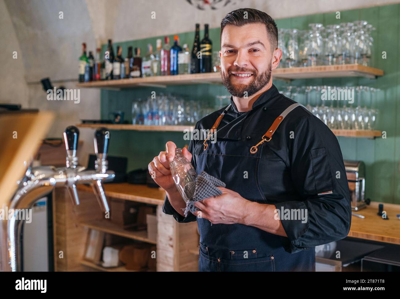Ritratto del barman barbarbato sorridente vestito in uniforme nera con un grembiule che strofina il bicchiere di birra al bancone del bar. Persone di successo, duro lavoro, Foto Stock