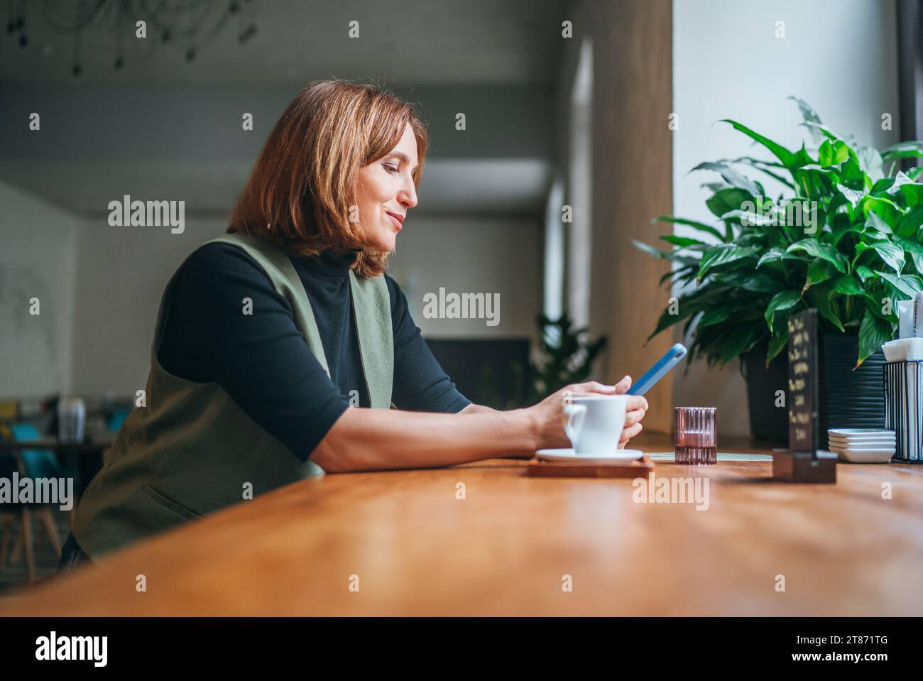 Ritratto di una bruna donna caucasica seduto al tavolo in un accogliente caffè con una piccola tazza di caffè e navigando in Internet utilizzando uno smartphone moderno. Techno moderno Foto Stock