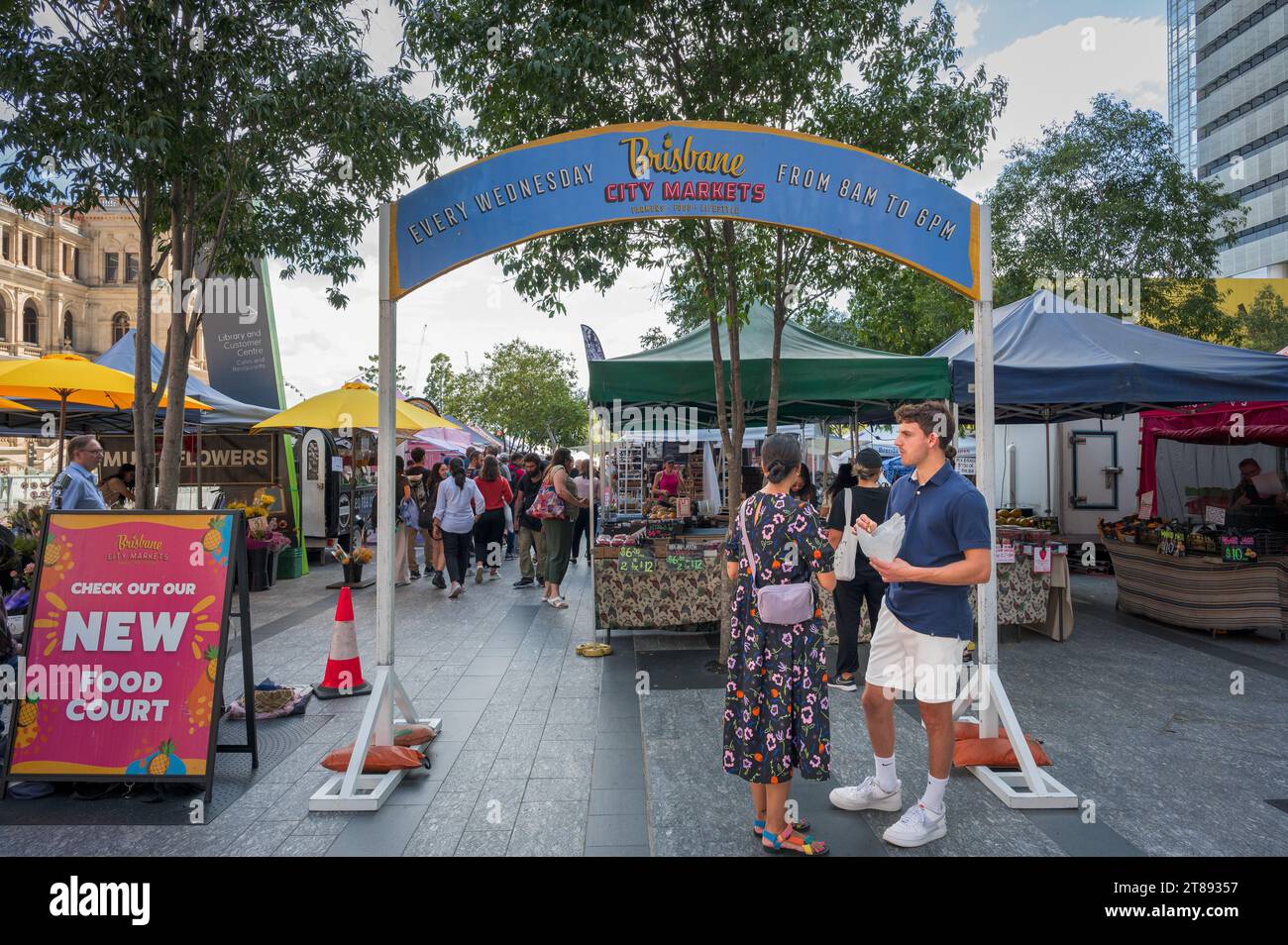Una giovane coppia si trova vicino al cartello dell'arco dei mercati di Brisbane, assaggiando alcuni dei piatti disponibili sia per la gente del posto che per i turisti. Foto Stock