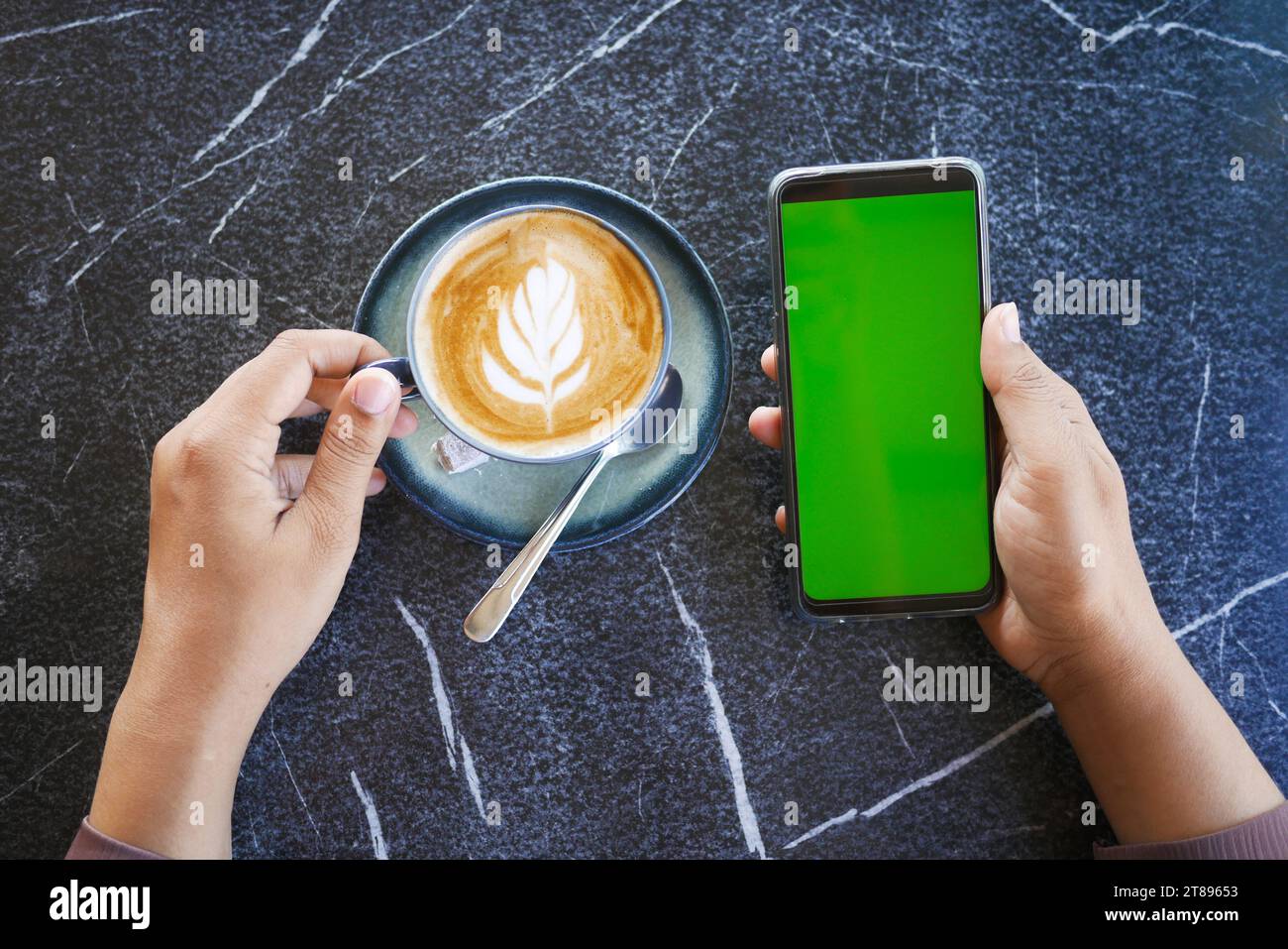 mano che regge uno smartphone con schermo verde e una tazza di caffè sul tavolo Foto Stock