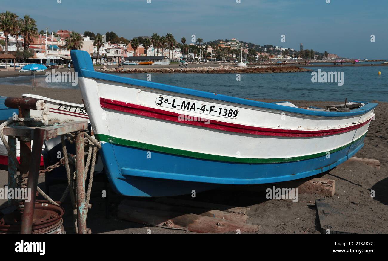 Villaggio di pescatori di Pedregalejo, Malaga, Andalusia: Tradizionale barca a remi in legno dipinto per la pesca si trova sulla sabbia; passeggiata sul lungomare alle spalle Foto Stock