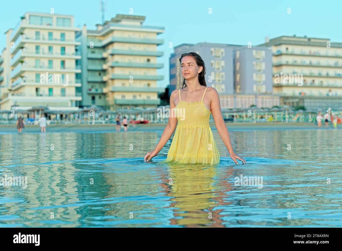 Giovane donna sulla spiaggia con un vestito giallo. Vacanze estive in spiaggia. La ragazza sta con le braccia incrociate dietro la testa Foto Stock