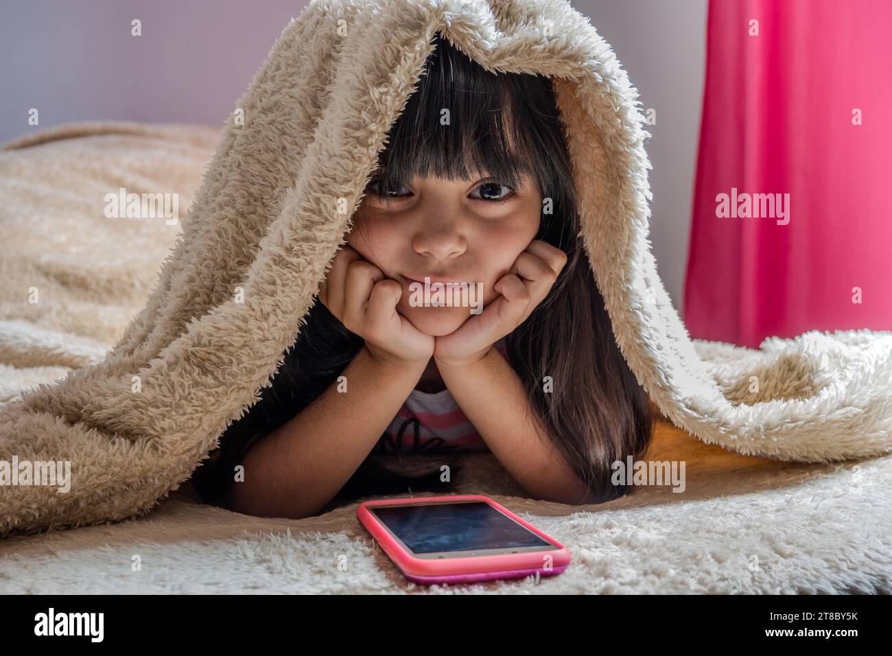Un bambino che utilizza uno smartphone sdraiato a letto, gioca, guarda video online, scorre lo schermo. Dipendenza da schermo per bambini. Stanza dei bambini. Foto Stock