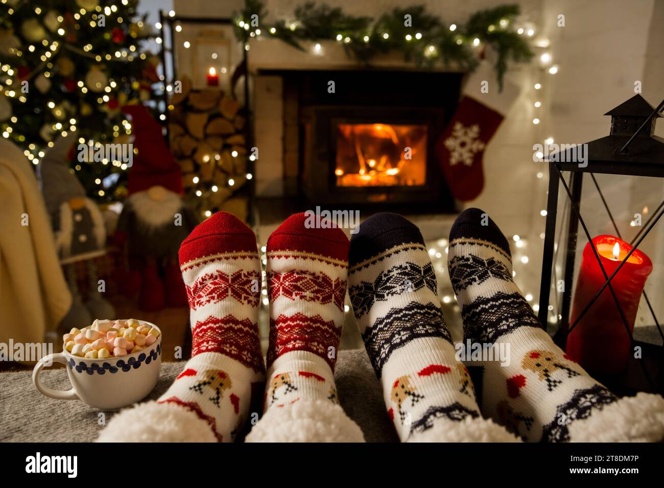 Coppia che si diverte davanti al caldo caminetto e si riscalda con calze di lana con ornamenti natalizi, albero di Natale e decorazioni di sfondo. Accogliente inverno, Natale e famiglia. Foto Stock