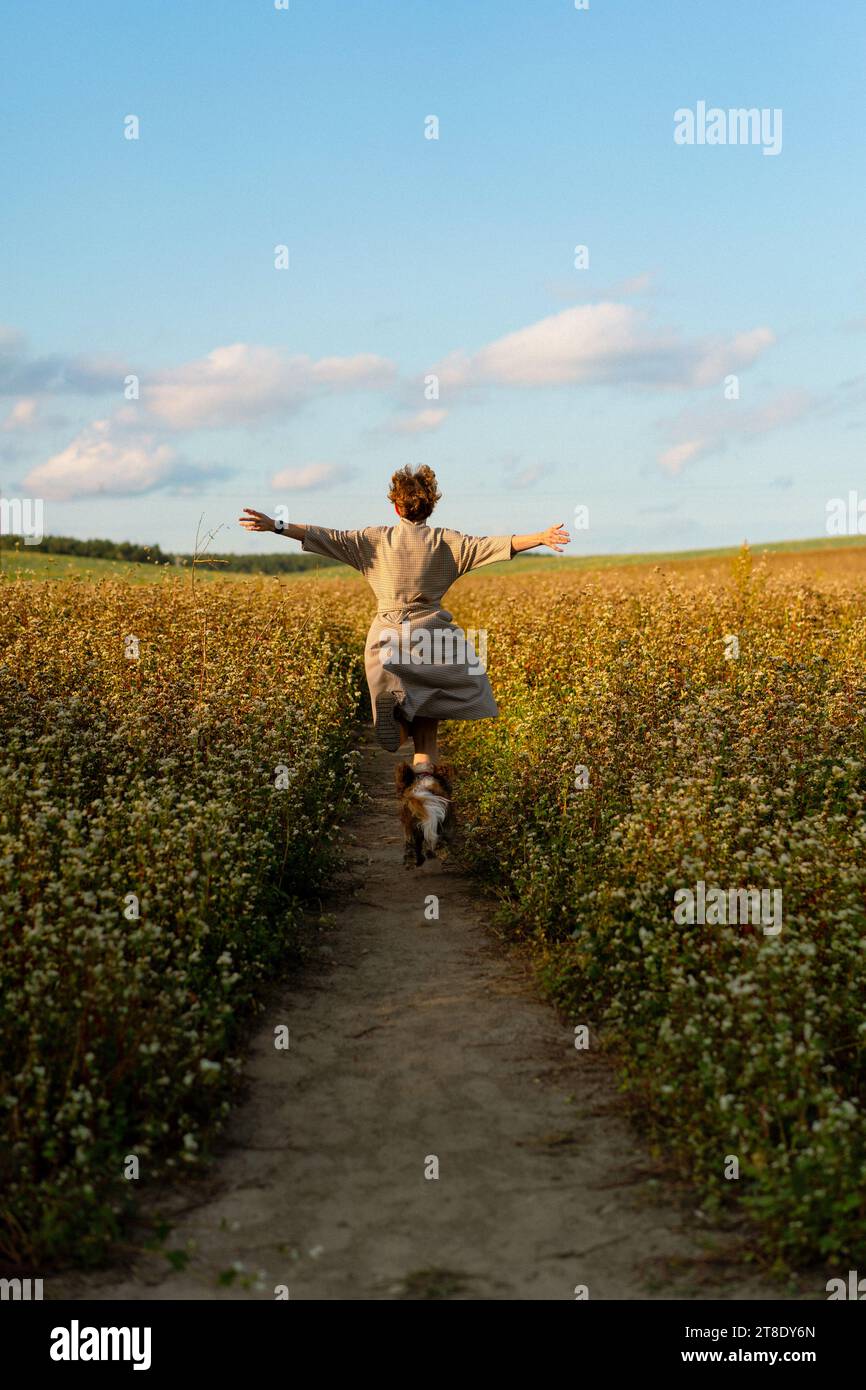 Una giovane donna con un cane corre in un campo giallo estivo al tramonto. Foto Stock