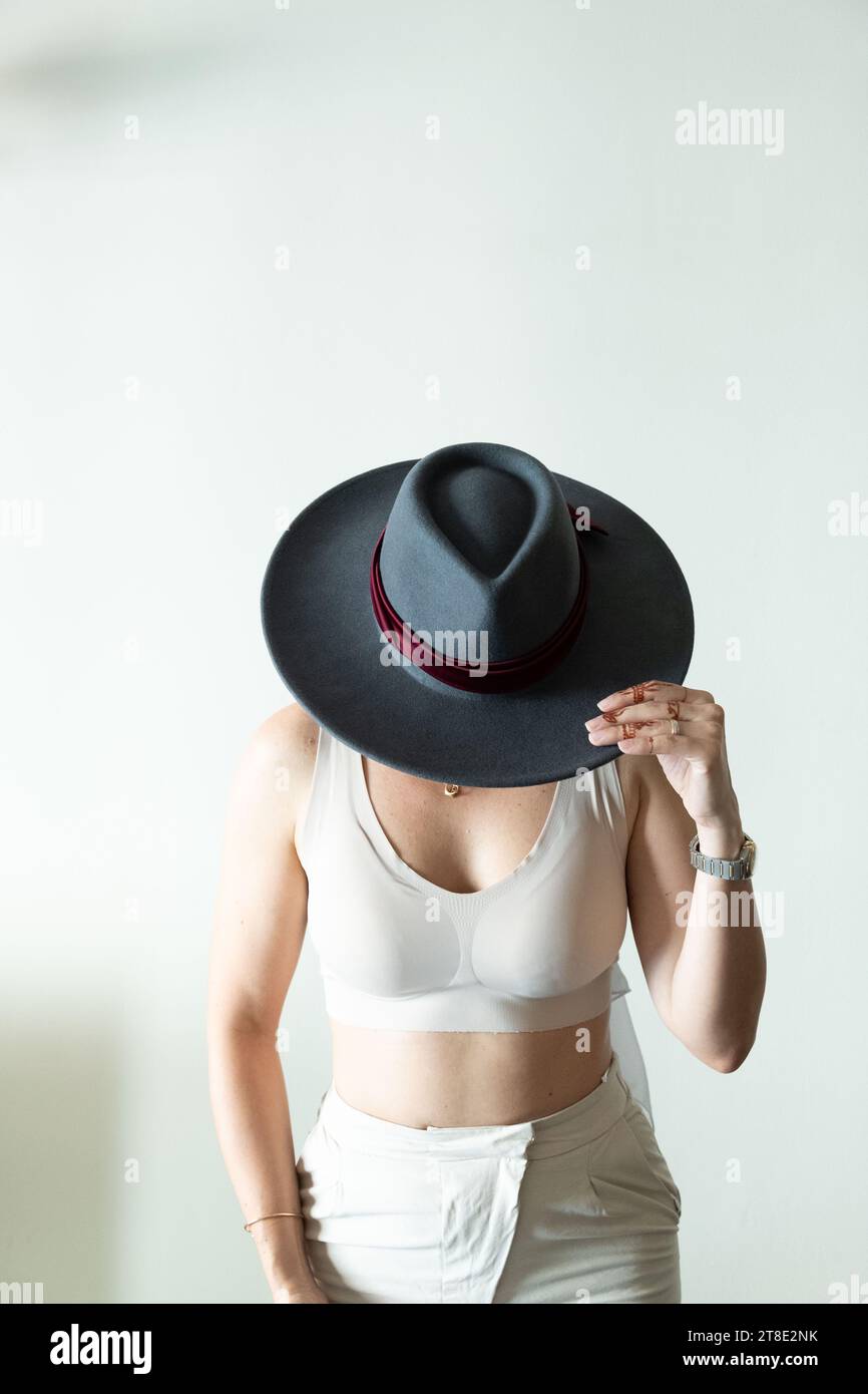 Donna vestita con colori neutri, indossa un elegante cappello grigio e posa su uno sfondo bianco Foto Stock