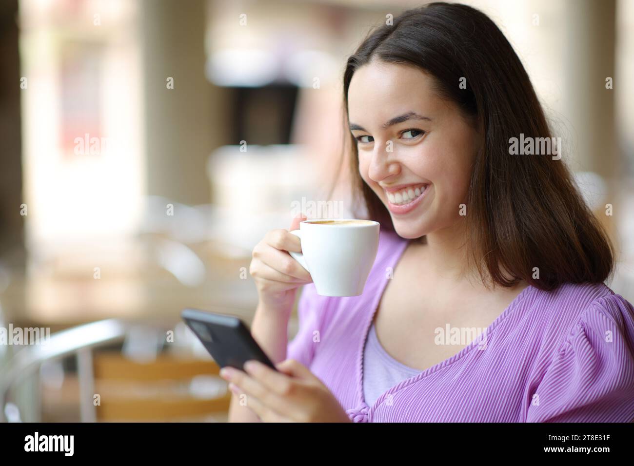 Una donna felice che guarda la macchina fotografica mentre tiene il telefono e il caffè in una terrazza del ristorante Foto Stock