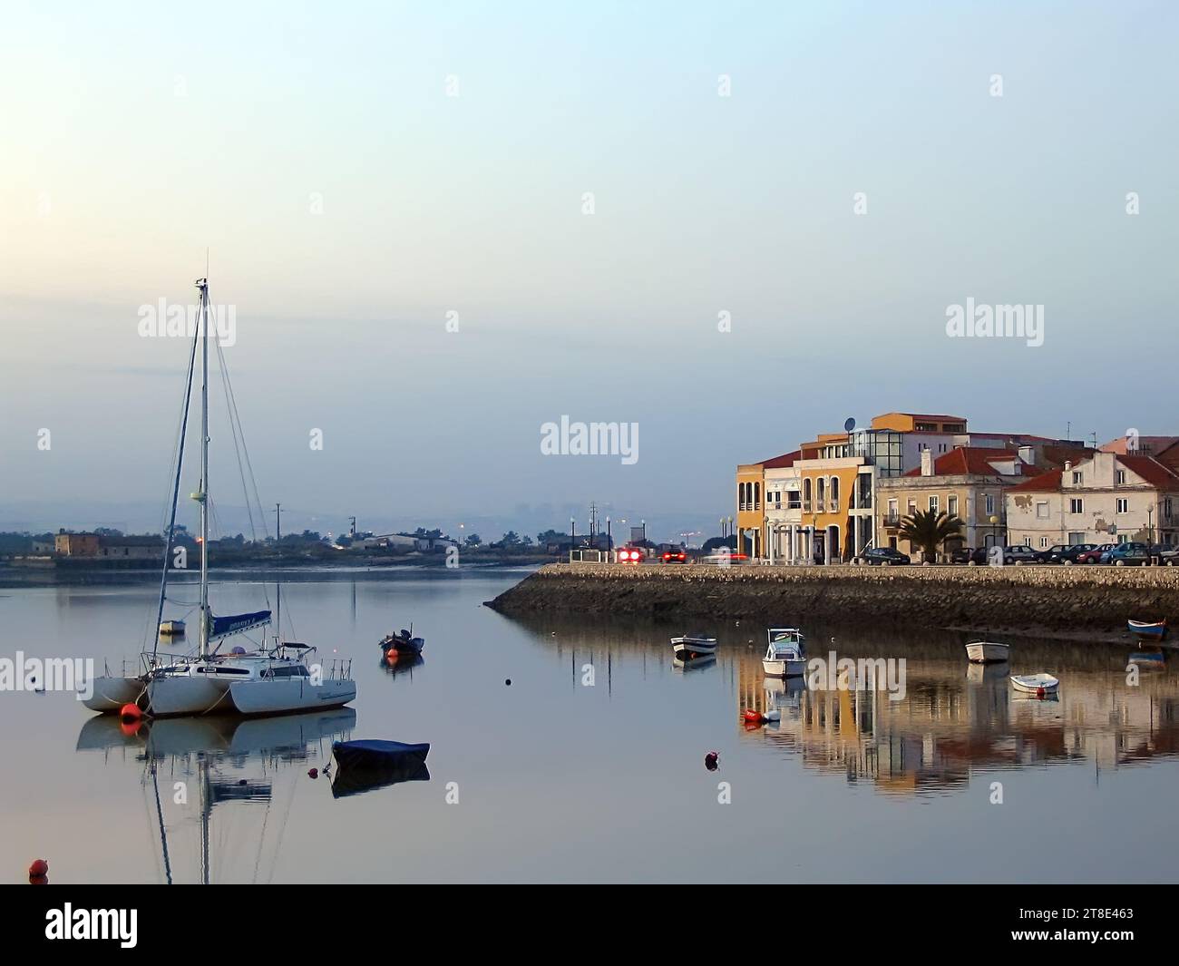 Vista della città di Seixal e della baia di Seixal con piccole barche e un trimarano a vela al crepuscolo, al tramonto o in serata. Setubal, Portogallo Foto Stock