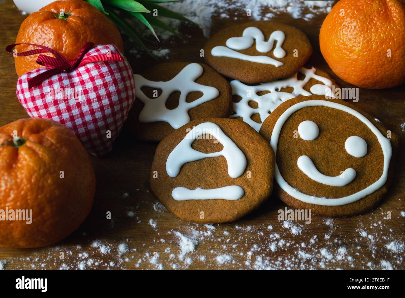 Biscotti dolci al pan di zenzero ricoperti di glassa bianca con vari motivi, circondati da mandarini e un cuore giocattolo su una tavola con farina Foto Stock