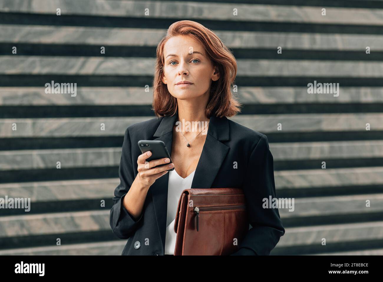 Donna d'affari di mezza età che tiene una cartella in pelle e uno smartphone e guarda la fotocamera mentre si trova all'aperto Foto Stock