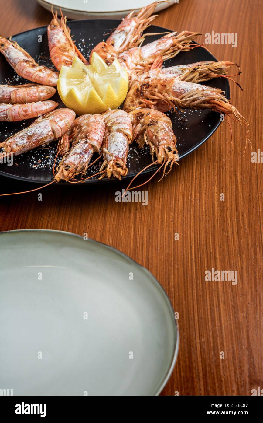 Foto verticale ravvicinata dei gamberi grigliati serviti con scaglie di limone e sale su un piatto nero e un tavolo di legno. Nessuna persona. Copia spazio. Gastronomia conce Foto Stock