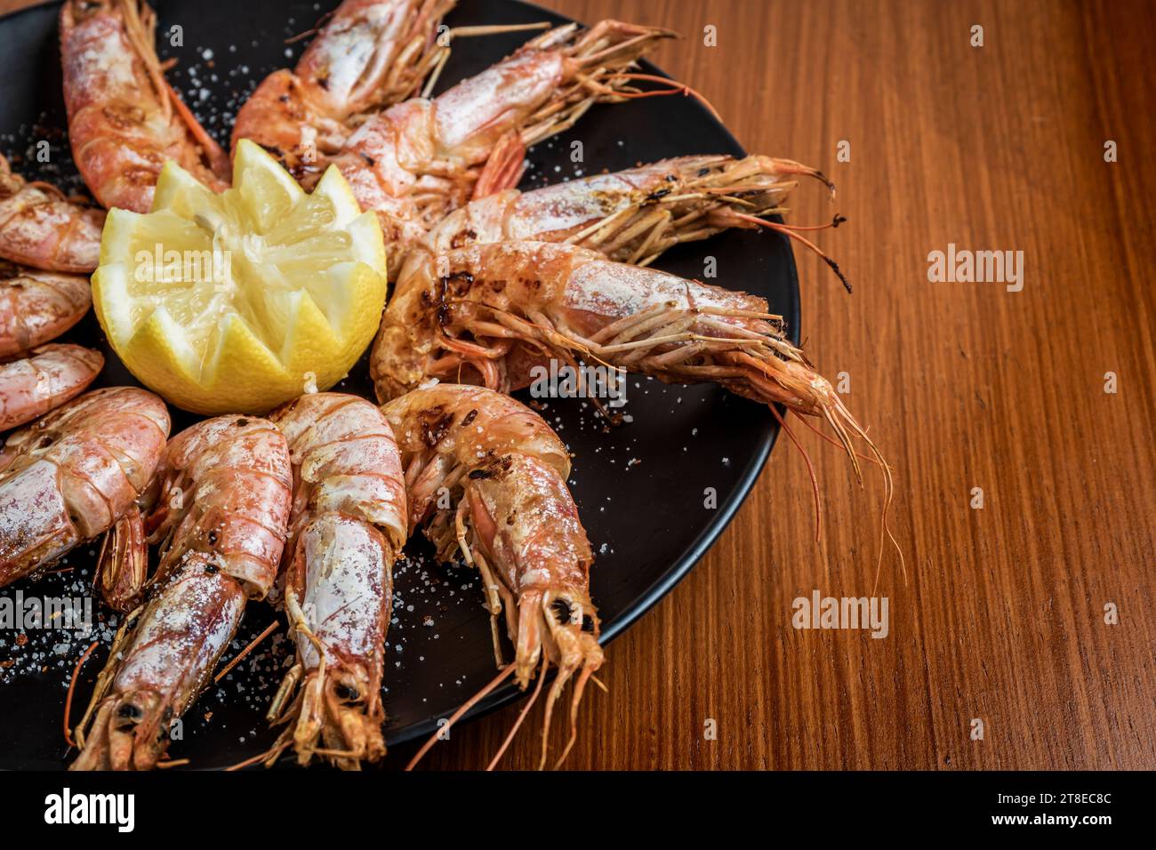 Foto orizzontale ravvicinata dei gamberi alla griglia serviti con scaglie di limone e sale su un piatto nero e un tavolo di legno. Nessuna persona. Copia spazio. Gastronomia con Foto Stock