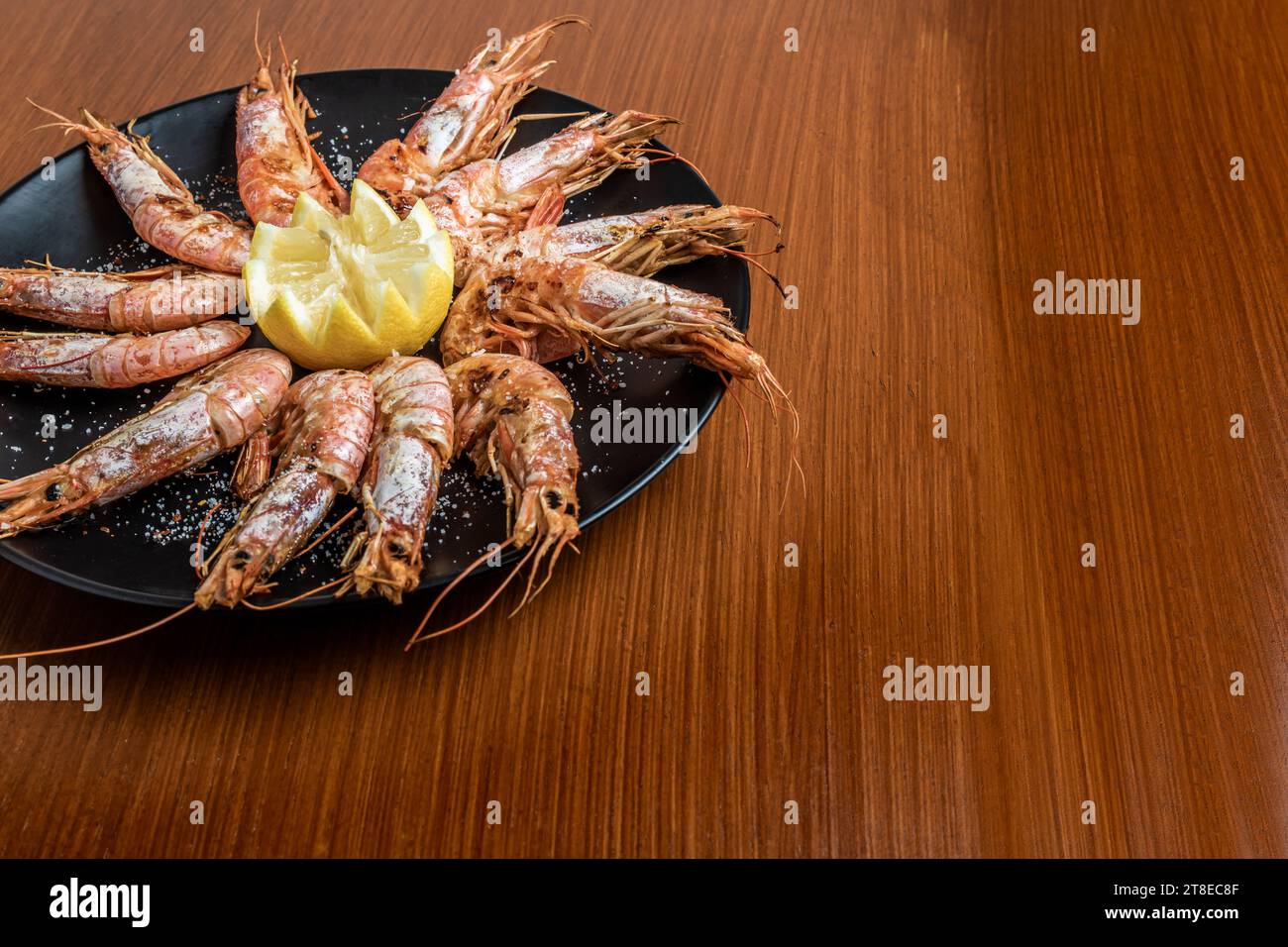 Foto orizzontale dei gamberi grigliati serviti con scaglie di limone e sale su un piatto nero e un tavolo di legno. Nessuna persona. Copia spazio. Concetto gastronomico. Foto Stock