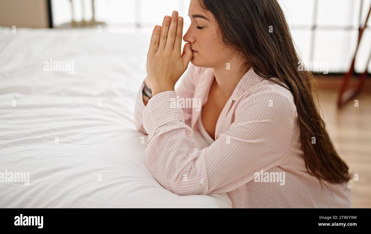 Giovane bella donna ispanica appoggiata sul letto che prega in camera da letto Foto Stock