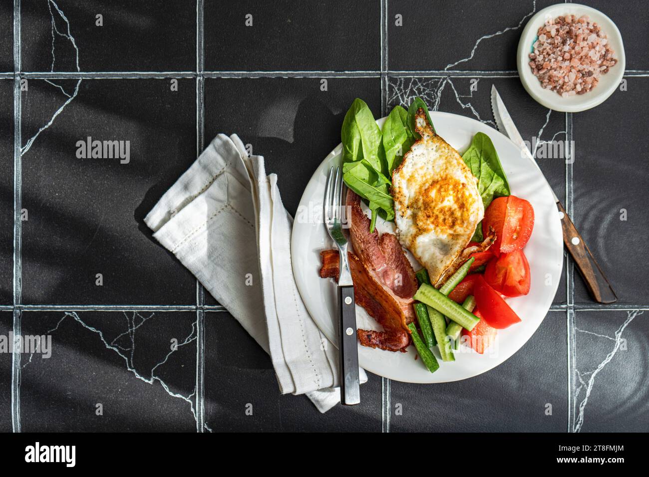 Vista dall'alto della colazione ben bilanciata con uova fritte, strisce di pancetta e verdure assortite come pomodoro, cetriolo e spinaci su un piatto bianco Foto Stock