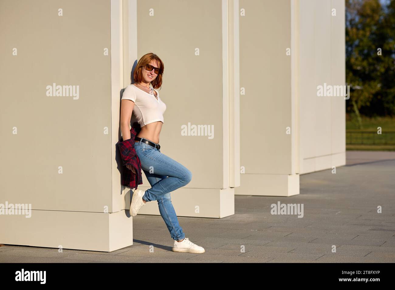 Una donna alla moda rossa che si gode una serata autunnale in città. Indossa una giacca rossa alla moda, una t-shirt bianca e jeans denim per un look audace Foto Stock