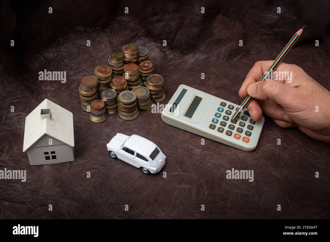 Mano che regge un pastello e calcola le tasse con la casa modello, l'auto e le pile multiple di monete su pelle marrone Foto Stock