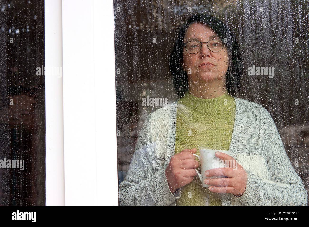 donna con una caffettiera che guarda fuori dalla finestra in un giorno di pioggia, perduta nel pensiero Foto Stock