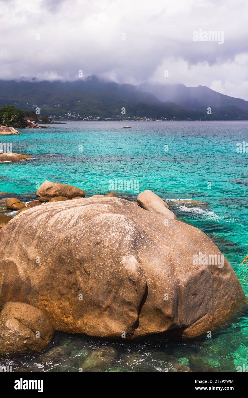 Vista do Mar Beach, isola di Mahe, Seychelles. Foto di paesaggio verticale con pietre costiere e acqua blu dell'oceano sotto il cielo nuvoloso in una giornata estiva Foto Stock