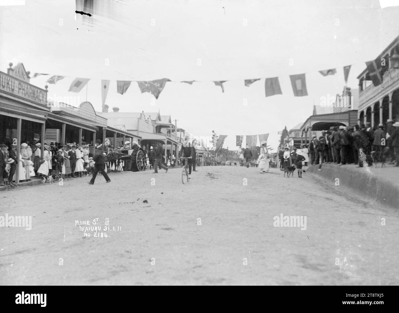 Queen Street, Waiuku, Queen Street, Waiuku, con corde di bandiere appese dall'altra parte della strada, e grandi gruppi di persone in piedi sotto le verande dei negozi. Fotografia scattata il 31 gennaio 1911 Foto Stock