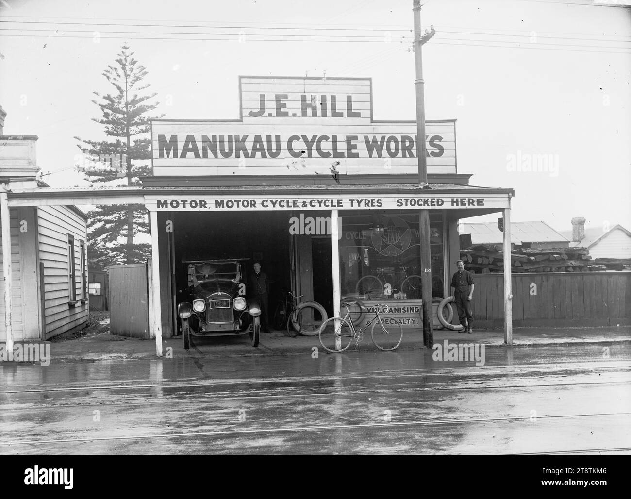J e Hill, Manukau Cycle Works, Onehunga, Vista della Manukau Cycle Works, Victoria Street, Onehunga (proprietario J e Hill) con un'auto parcheggiata nell'ingresso del garage sul lato sinistro del negozio. Una bicicletta è appoggiata contro il supporto della veranda di fronte al negozio, e un uomo si trova sulla destra a guardare il fotografo. La pubblicità sul fronte del negozio recita "pneumatici per motori, motocicli e biciclette qui forniti" e "vulcanizzare una specialità". Un pino Norfolk cresce dietro la proprietà accanto. Il legno è impilato dietro la recinzione della proprietà vicina Foto Stock
