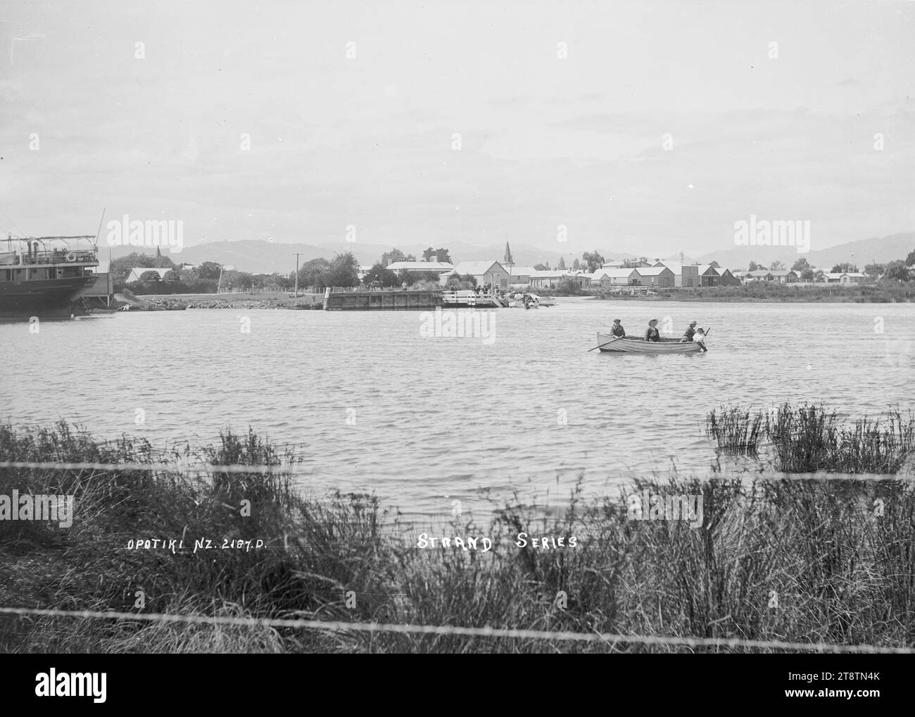 Vista del fiume Waioeka e di Opotiki, vista del fiume Waioeka che guarda a sud dalla riva del fiume con la cittadina di Opotiki in lontananza. Il molo è a media distanza e una barca a remi con quattro persone a bordo è in primo piano. A sinistra si trova la poppa di una grande nave marittima. all'inizio degli anni '1900 Foto Stock