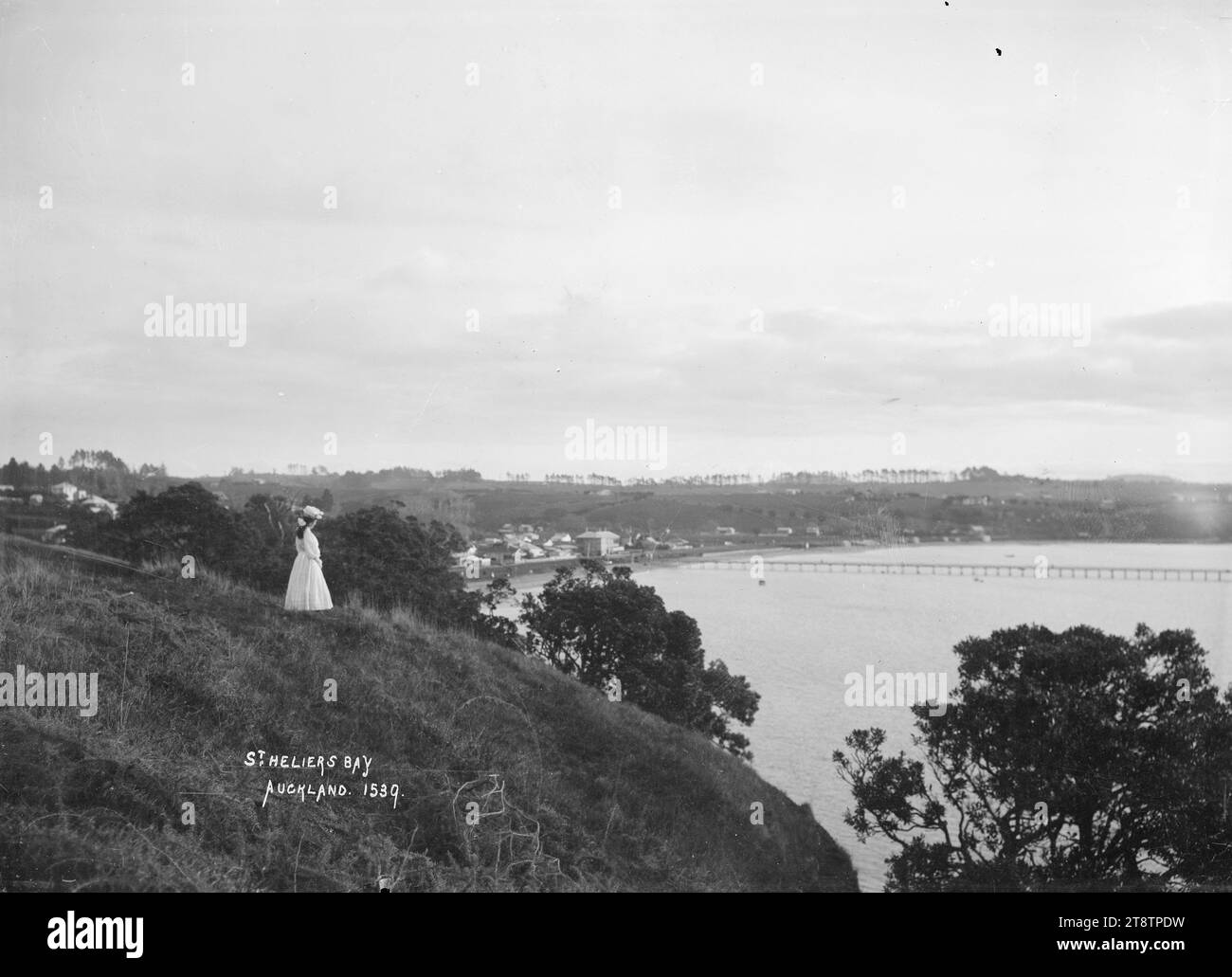 St Heliers Bay, St Heliers, Auckland, nuova Zelanda da Achilles Point, Vista presa dalle vicinanze di Achilles Point guardando a sud-ovest verso St Heliers Bay. St Heliers Bay Hotel e molo a media distanza. Una giovane donna è in piedi vicino alla cima della scogliera e guarda verso la baia. Fotografato tra il 1910 e il 1930 Foto Stock