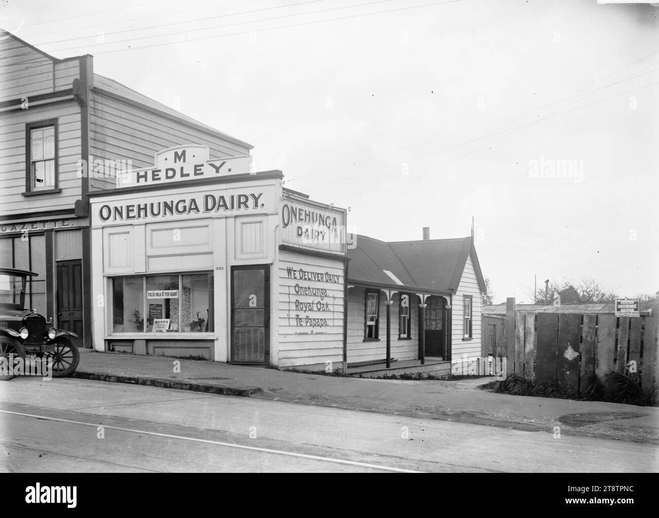 Onehunga Dairy, Queen Street, Onehunga, Vista di Onehunga Dairy, 223 Queen Street, Onehunga (proprietario M Hedley) con un unico piano che si trova dietro. "Consegniamo ogni giorno - Onehunga, Royal Oak, te Papapa' è scritto sul lato del negozio. I cartelli nella vetrina del negozio pubblicizzano "latte fresco 6d per quart" e "pane fatto con latte del Kent". Accanto c'è un'azienda di stampa. Sulla recinzione a destra c'è un cartello "in vendita" (Horne, l'uomo di terra). Un'auto è parcheggiata sul marciapiede di fronte al negozio, CA 1920s Foto Stock