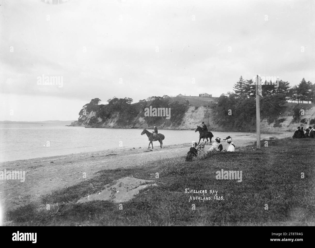 St Heliers Bay, St Heliers, Auckland, nuova Zelanda, Vista dell'estremità orientale della St Heliers Bay guardando verso Archilles Point. Gruppi di persone sono seduti nell'area erbosa vicino alla spiaggia. Due uomini cavalcano cavalli sulla sabbia nelle vicinanze. Fotografato tra il 1910 e il 1930 Foto Stock