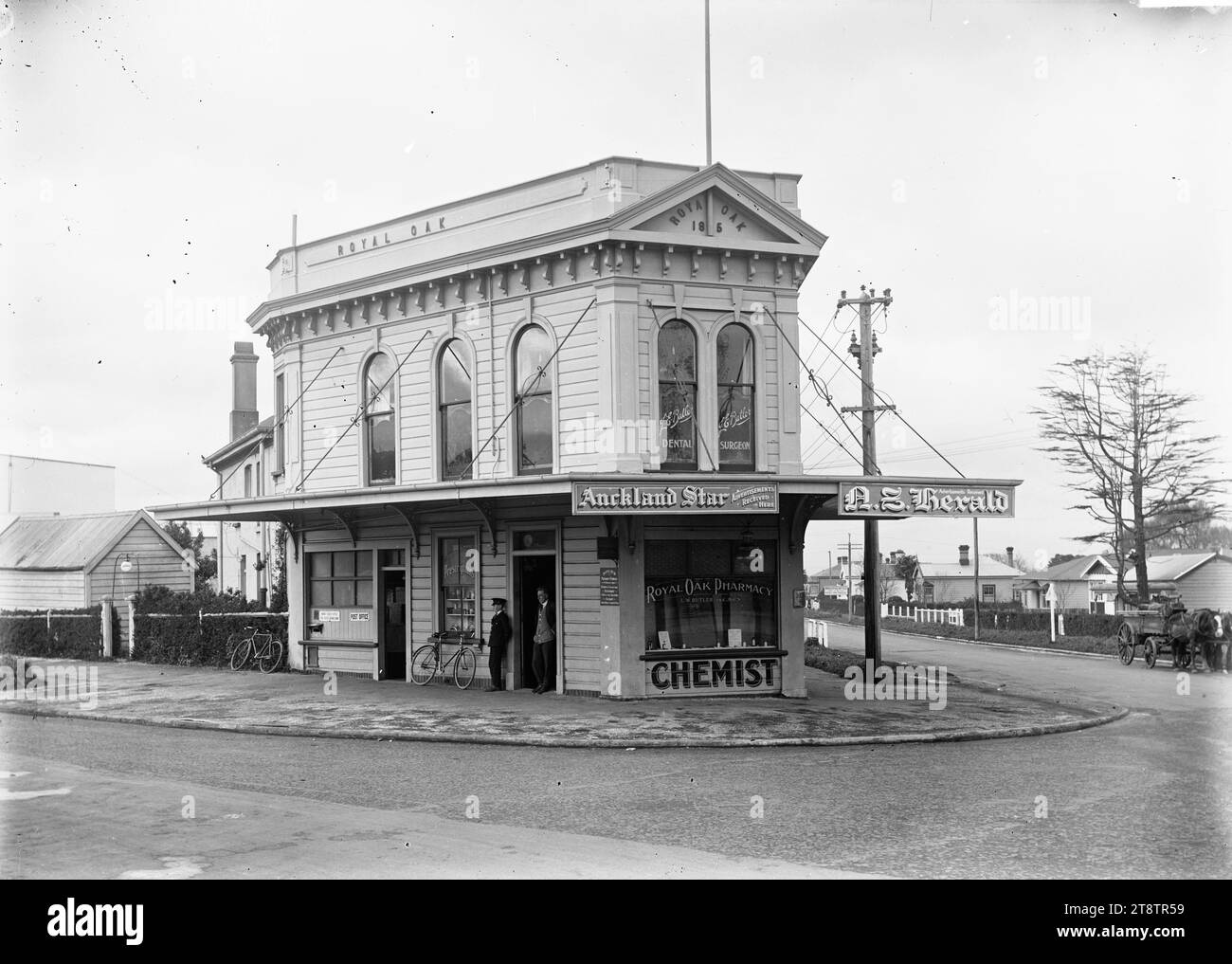 Business all'angolo tra Campbell Road e Mt Smart Road, Royal Oak, Auckland, nuova Zelanda, vista dell'edificio all'angolo tra Mt Smart Road e Campbell Road, Royal Oak. La Royal Oak Pharmacy e un ufficio postale occupano le aree al piano inferiore e J e Butler, chirurgo dentale, dispone di camere al piano superiore. Due uomini possono essere visti su una porta (uno è un postino) e ci sono due biciclette nelle vicinanze. Una casa e un carro sono visibili sulla strada a destra. Si possono vedere i cartelli Auckland, New Zealand Star e NZ Herald. L'edificio sul retro con un camino è l'originale hotel Royal Oak costruito nel 1853 Foto Stock