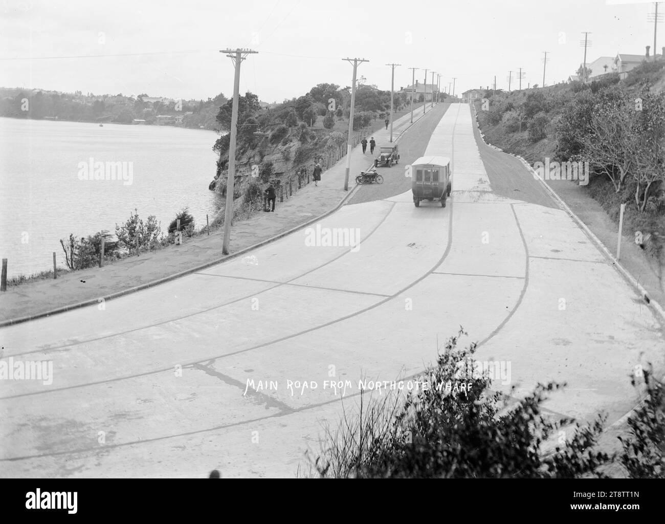 Strada principale da Northcote Wharf, Auckland, nuova Zelanda, vista su Queen Street dal Northcote Wharf End, mostra un autobus che viaggia lontano dal molo, e una moto e un'auto parcheggiate sul lato della strada. Diverse persone possono essere viste camminare sul sentiero accanto alla strada di cemento. Little Shoal Bay può essere vista sul lato destro. ca 1920 Foto Stock