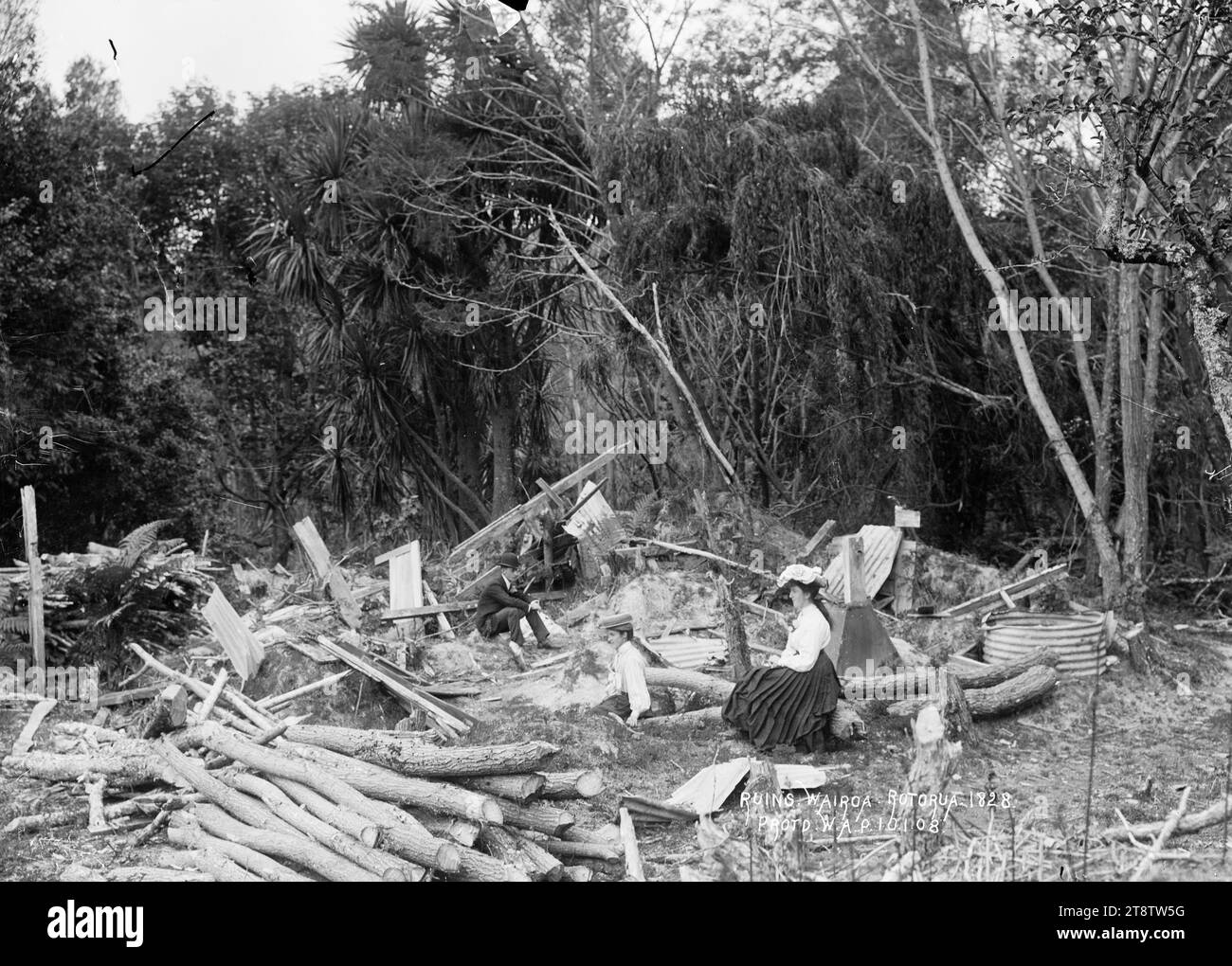 Rovine a te Wairoa, vicino a Rotorua, veduta di un uomo e due donne sedute tra legname, ferro ondulato e tronchi nel sito di edifici demoliti, sullo sfondo di un cespuglio nativo. Si possono vedere i resti di un serbatoio d'acqua. Il 10 gennaio 1908 Foto Stock