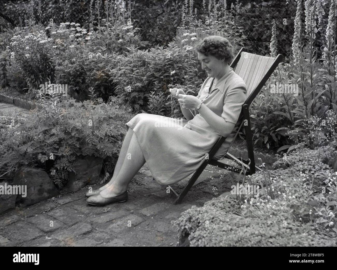 Anni '1950, storica, una giovane donna seduta su una sdraio su un sentiero in un giardino di cottage con un ago e un filo da cucito, Inghilterra, Regno Unito. Foto Stock